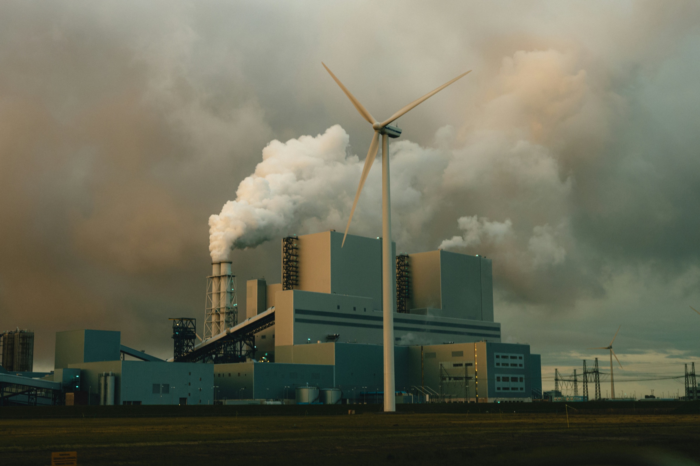 A wind turbine in front of an industrial power plant under heavy clouds