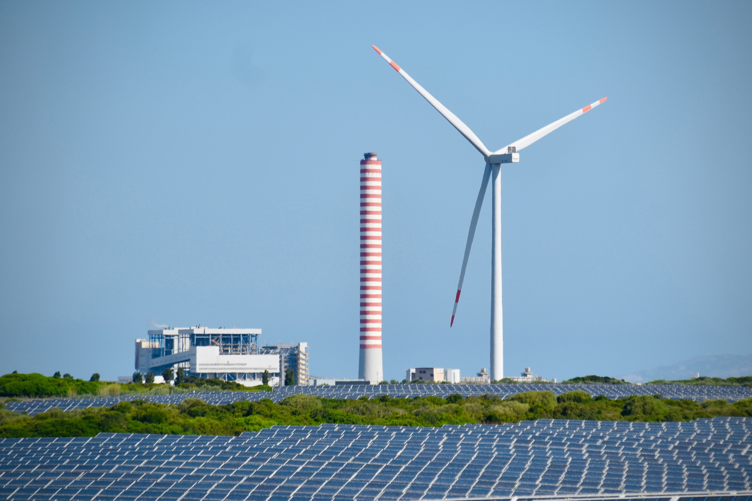 A wind turbine and solar panels beside a power plant under a clear blue sky